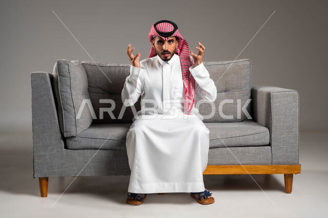 ايماءات التوتر والانزعاج وعدم تحمل الطفح الجلدي, بورتريه لشاب عربي خليجي سعودي يرتدي الثوب والشماغ التقليدي يجلس على الكنبة ويرفع يديه للأعلى, المشاكل الصحية وتهيجات الجلد, بثور منتشرة على بشرة الوجه, الإصابة الجلدية وفرط التحسس, خلفية رمادية