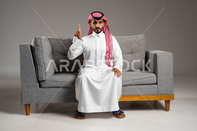 الطفح الجلدي والأمراض المعدية, القيام بالتحاليل اللازمة, بورتريه لشاب عربي خليجي سعودي يرتدي الثوب والشماغ التقليدي يجلس على الكنبة ويرفع اصبعه السبابة للأعلى, المشاكل الصحية وتهيجات الجلد, بثور منتشرة على بشرة الوجه, الإصابة الجلدية وفرط التحسس, خلفية رمادية