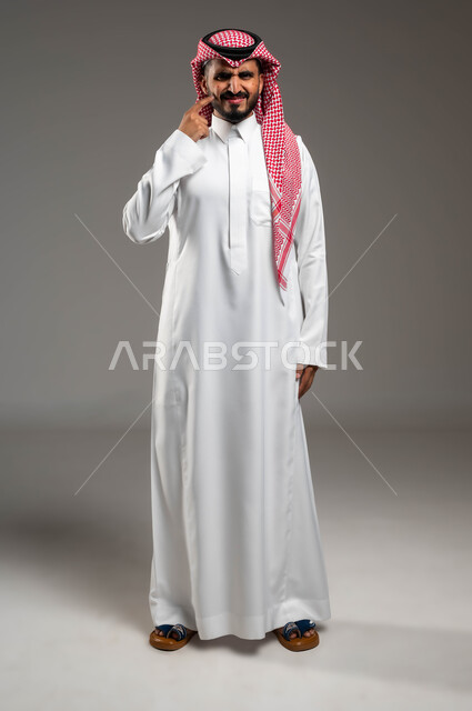 الوقوف والإشارة إلى مكان الإصابة, بثور منتشرة على بشرة الوجه, بورتريه لرجل عربي خليجي سعودي يرتدي الثوب التقليدي والشماغ يقف بإيماءات الإنزعاج والحزن, الإصابة بالحساسية الجلدية, المشاكل الصحية والتهجيات والشعور بالألم, صورة الجسم بالطول الكامل, خلفية رمادية