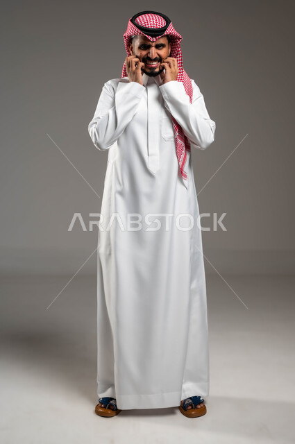 المشاكل الصحية والإصابة بالحساسية الجلدية, الأمراض والتهجيات وفرط التحسس, الشعور بالآلام والأوجاع, بورتريه لرجل عربي خليجي سعودي يرتدي الثوب والشماغ التقليدي يقف بإيماءات الألم, بثور منتشرة على بشرة الوجه, صورة الجسم بالكامل, خلفية رمادية