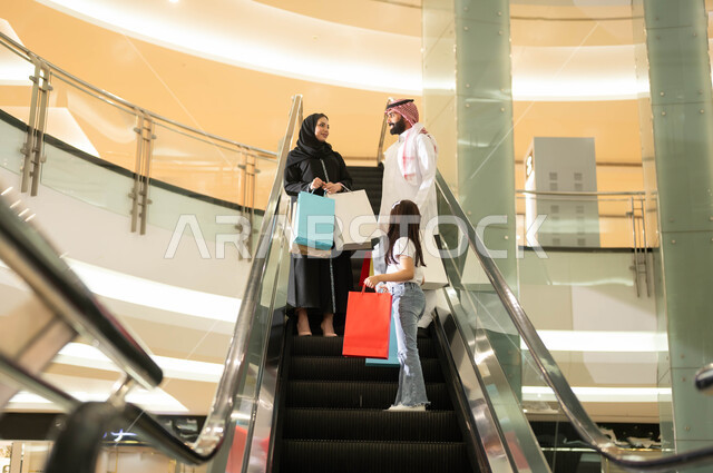 الانتقال والنزول باستخدام السلم الكهربائي, عائلة عربية خليجية سعودية تحمل أكياس ورقية ملونة تتجول في مركز التسوق، الاستمتاع بالشراء وانتقاء المستلزمات والهدايا، الاستفادة من العروض والخصومات في المول التجاري، مفهوم الأسرة السعيدة