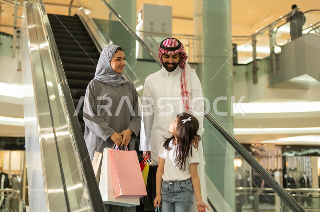 الانتقال والنزول باستخدام السلم الكهربائي, عائلة عربية خليجية سعودية تحمل أكياس ورقية ملونة تتجول في مركز التسوق، الاستمتاع بالشراء وانتقاء المستلزمات والهدايا، الاستفادة من العروض والخصومات في المول التجاري، مفهوم الأسرة السعيدة