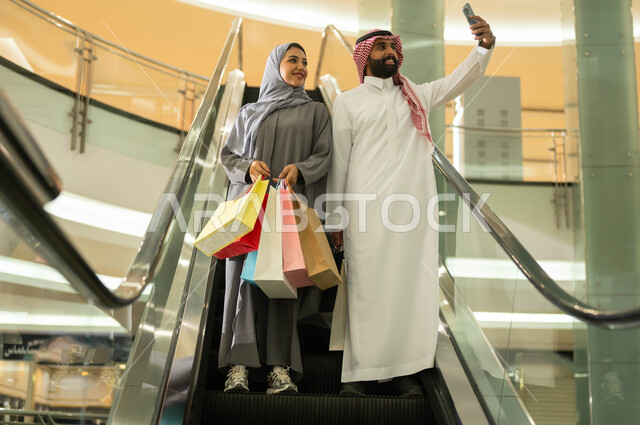 النزول على السلم الكهربائي والتقاط صور سيلفي تذكارية باستخدام الهاتف الجوال, اختيار وشراء المستلزمات والهدايا, قضاء وقت ممتع برفقة الأسرة, زوجان عربيان سعوديان خليجيان يقومان بالتجول في إحدى المولات التجاريه, متعة التسوق في الأسواق السعوديه