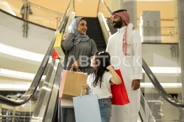 النزول إلى الطابق الآخر باستخدام السلم الكهربائي, مفهوم الأسرة السعيدة، شراء واقتناء المستلزمات من المول، عائلة عربية خليجية سعودية يحملون أكياس التسوق الملونة, التجول والتسوق في إحدى المولات التجارية في المملكة