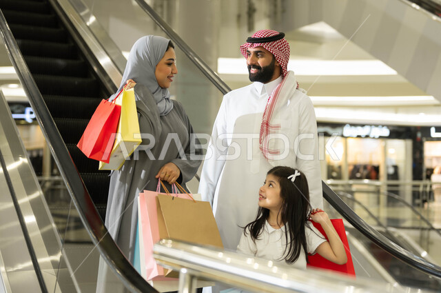 شراء واقتناء المستلزمات من المول، النزول والانتقال إلى الطابق الآخر باستخدام السلم الكهربائي, عائلة عربية خليجية سعودية يحملون أكياس التسوق الملونة, التجول والتسوق في إحدى المولات التجارية في المملكة, مفهوم الأسرة السعيدة