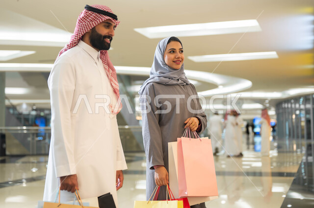 قضاء وقت ممتع في إحدى المولات التجارية في المملكة, مشاركة الزوجين في اختيارات التسوق وتبادل الآراء, رجل وامرأة عربيان سعوديان خليجيان يحملان أكياس التسوق الملونة, الاستفاده من العروض والتخفيضات الموسمية