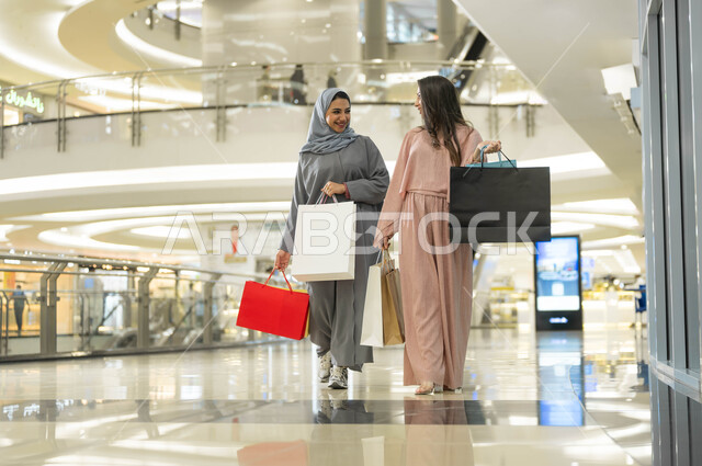 متعة مشاركة الأصدقاء في اختيار الهدايا من المراكز التجارية، الأثر الإيجابي للتسوق على النساء، إمرأتان عربيتان خليجيتان سعوديتان مبتسمتان تحملان الأكياس الورقية تقومان بالتبضع في المول، تبادل الاحاديث والاخبار اثناء التسوق، الاستفادة من حسومات الجمعة البيضاء
