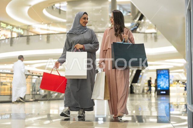 تبادل الاحاديث والاخبار اثناء التسوق، الاستفادة من حسومات الجمعة البيضاء، متعة مشاركة الأصدقاء في اختيار الهدايا من المراكز التجارية، الأثر الإيجابي للتسوق على النساء، إمرأتان عربيتان خليجيتان سعوديتان مبتسمتان تحملان الأكياس الورقية تقومان بالتبضع في المول