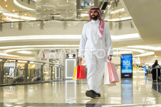 Expressions of joy and happiness, White Friday discounts and offers in Saudi Arabia, a smiling Saudi Gulf Arab man wearing a shemagh and traditional dress, carrying colorful paper bags in his hand, walking around inside the mall, the joy of shopping and purchasing supplies and gifts from stores and malls.