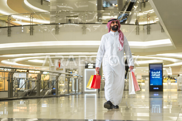 إيماءات السرور والسعادة، حسومات وعروض الجمعة البيضاء في السعودية، رجل عربي خليجي سعودي مبتسم يرتدي الشماغ والثوب التقليدي يحمل بيده أكياس ورقية ملونة يقوم بالتجول داخل المول التجاري، متعة التسوق واقتناء المستلزمات والهدايا من المحلات والمراكز التجاريه