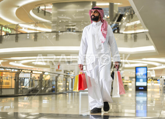 Expressions of joy and happiness, White Friday discounts and offers in Saudi Arabia, a smiling Saudi Gulf Arab man wearing a shemagh and traditional dress, carrying colorful paper bags in his hand, walking around inside the mall, the joy of shopping and purchasing supplies and gifts from stores and malls.
