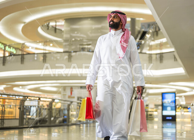 إيماءات السرور والسعادة، حسومات وعروض الجمعة البيضاء في السعودية، رجل عربي خليجي سعودي مبتسم يرتدي الشماغ والثوب التقليدي يحمل بيده أكياس ورقية ملونة يقوم بالتجول داخل المول التجاري، متعة التسوق واقتناء المستلزمات والهدايا من المحلات والمراكز التجاريه