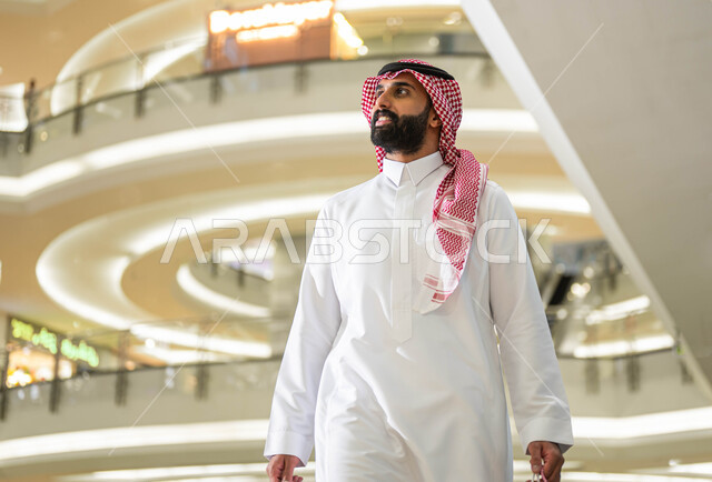 Buying supplies and gifts from stores and malls, a close-up picture of a smiling Saudi Gulf Arab man wearing a shemagh and traditional thobe, walking around inside the mall, gestures and expressions of pleasure and happiness, taking advantage of the White Friday discounts and offers in Saudi Arabia.