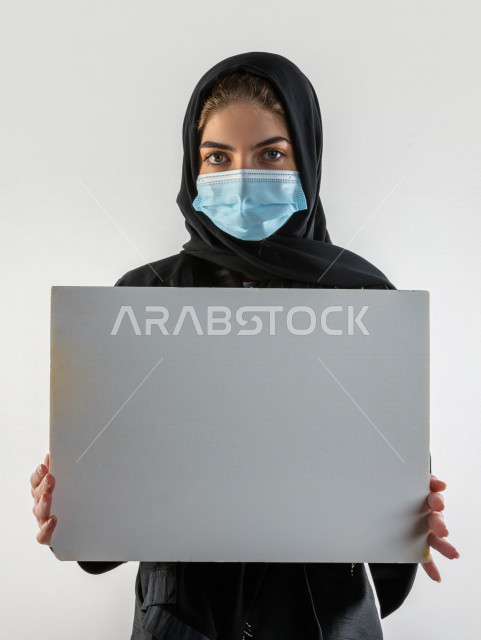 بورتريه لشابة سعودية تضع الكمامة ،  تحمل بيدها لوح ابيض فارغ ، اتباع اجراءات السلامة بوضع الكمامة بشكل صحيح ، تجنب انتشار العدوى ، خلفية بيضاء