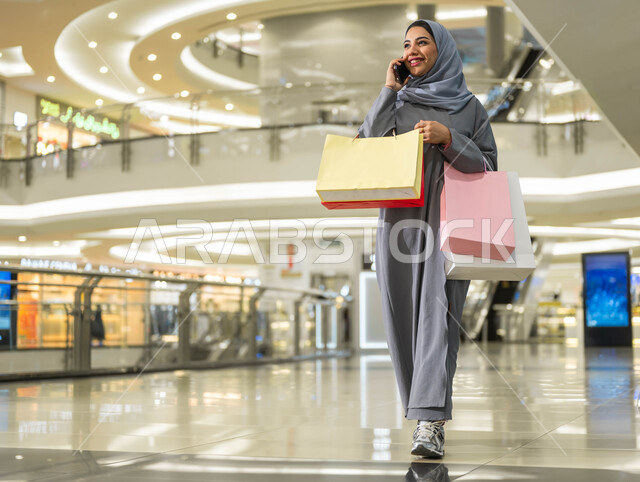 استخدام الهاتف المحمول أثناء التسوق داخل المول، الترويج لماركات عالمية فاخرة، الاستفادة من العروض والتخفيضات الموسمية في الأسواق، اقتناء الهدايا والمستلزمات، بورتريه لامرأة عربية خليجية سعودية محجبة ترتدي العباءة تقوم بإجراء مكالمة هاتفية