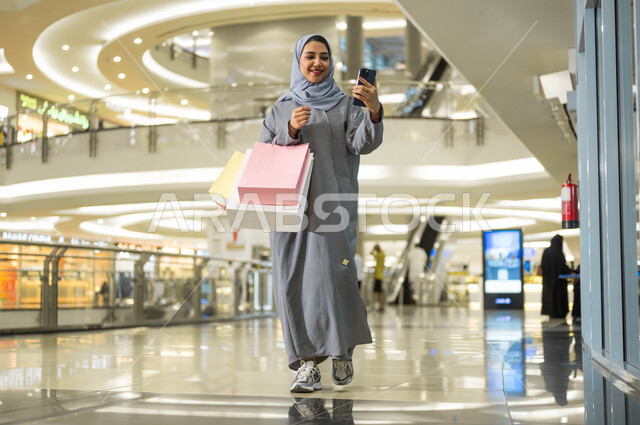 الترويج لماركات عالمية فاخرة، الاستفادة من العروض والتخفيضات الموسمية في الأسواق، اقتناء الهدايا والمستلزمات، بورتريه لامرأة عربية خليجية سعودية محجبة ترتدي العباءة تقوم بالتقاط صور السيلفي باستخدام الهاتف المحمول أثناء التسوق داخل المول