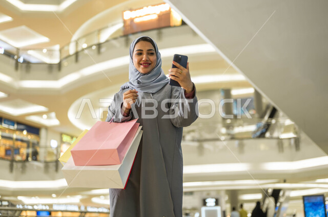 الترويج لماركات عالمية فاخرة، الاستفادة من العروض والتخفيضات الموسمية في الأسواق، اقتناء الهدايا والمستلزمات، بورتريه لامرأة عربية خليجية سعودية محجبة ترتدي العباءة تقوم بالتقاط صور السيلفي باستخدام الهاتف المحمول أثناء التسوق داخل المول