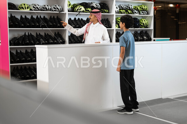 استعارة الزلاجات وخوذة الحماية من الاستقبال عند مضمار التزلج، موظف عربي خليجي سعودي يقوم بمساعدة ولد في اختيار الأدوات اللازمة لممارسة هواية التزلج في ساحات الجليد في أحد الأماكن الترفيهيه في السعودية، الاستمتاع في قضاء أوقات الفراغ في العطلة الصيفية