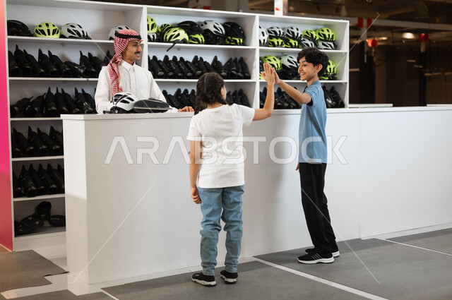 Raising hands with gestures of agreement, preparing to begin challenges and adventures, a picture from behind of a Saudi Gulf Arab boy and girl borrowing skates and a safety helmet from reception at the skating track in one of the entertainment centers in Saudi Arabia, practicing favorite hobbies in the skating rinks.