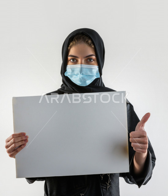 بورتريه لشابة سعودية تضع الكمامة ،  تحمل بيدها لوح ابيض فارغ ، اتباع اجراءات السلامة بوضع الكمامة بشكل صحيح ، تجنب انتشار العدوى ، خلفية بيضاء