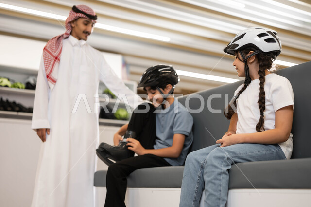 قضاء وقت ممتع أحد الأماكن الترفيهيه في السعودية، تطوير المهارات الرياضية، ايماءات الحماس لبدء السباقات، اللعب في ساحات التزلج، فتاة و ولد عربي خليجي سعودي يقوم بارتداء الزلاجات و خوذة الحماية، موظف سعودي يقوم بتبادل بتشجيع الأطفال على التحدي والمنافسة