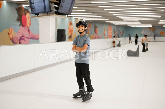 العمل على تطوير المهارات الرياضية، ولد عربي خليجي سعودي مبتسم يرتدي خوذه الحماية يقف بأيدي متقاطعة، ممارسة هواية التزلج على الجليد في إحدى الأماكن الترفيهيه في السعودية، قضاء عطلة صيفية ممتعه ،النظر الى الكاميرا بإيماءات السرور