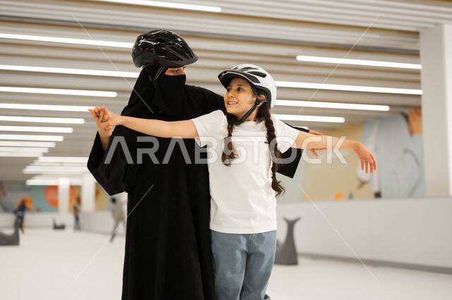 قضاء أوقات الفراغ في إحدى الأماكن الترفيهيه، تعليم الابناء على التزلج في ساحات الجليد ،اجراء تجارب مميزة، مفهوم المحبة و الترابط الأسري، الاهتمام في نفسية الأولاد ومنحهم الطاقة الايجابية، امرأة عربية خليجية سعودية محجبة تمسك بأيدي ابنتها لتساعدها باللعب