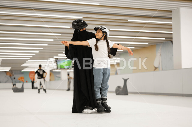 تعليم الابناء على التزلج في ساحات الجليد ،اجراء تجارب مميزة، مفهوم المحبة و الترابط الأسري، الاهتمام في نفسية الأولاد ومنحهم الطاقة الايجابية، قضاء أوقات ممتعه في إحدى الأماكن الترفيهيه، امرأة عربية خليجية سعودية محجبة تمسك بأيدي ابنتها لتساعدها باللعب