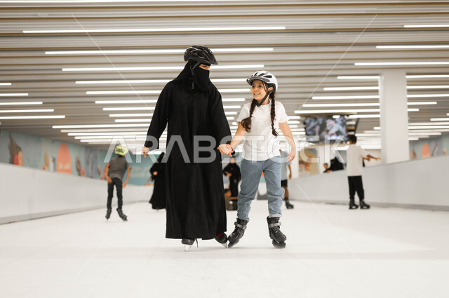 اجراء تجارب مميزة و مثيرة، مفهوم المحبة و الترابط الأسري، الاهتمام في نفسية الأولاد ومنحهم الطاقة الايجابية، قضاء أوقات ممتعه في إحدى الأماكن الترفيهيه المسلية مع العائلة، امرأة عربية خليجية سعودية محجبة تمسك يد ابنتها و تمارس لعبة التزلج في ساحات الجليد