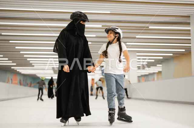 The concept of love and family cohesion, paying attention to the psychology of children and giving them positive energy, spending enjoyable times in one of the entertaining entertainment places with the family, conducting exciting and distinctive experiences, a veiled Saudi Arabian Gulf woman holding her daughter’s hand and playing skating in the ice rinks.