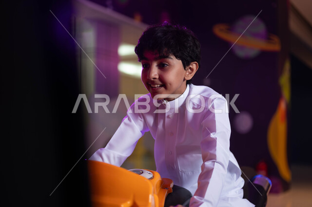 العاب الاركيد الالكترونيه المتطورة، الاستمتاع بقضاء الاجازة الصيفيه في احدى الصالات في الأماكن الترفيهية في السعودية ،ايماءات الاندماج و التركيز، مفهوم المنافسة والتحدي، صورة مقربة لولد عربي خليجي سعودي مبتسم يرتدي الزي التقليدي و يقوم بركوب الدباب الكهربائي