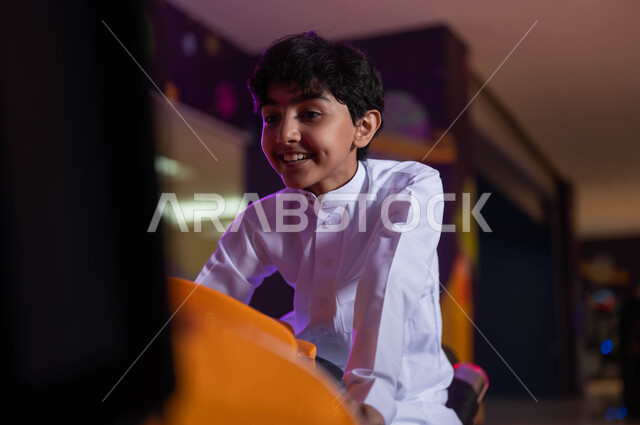 الاستمتاع بقضاء الاجازة الصيفيه في احدى الصالات في الأماكن الترفيهية في السعودية ،ايماءات الفرح والسرور، مفهوم المنافسة والتحدي، صورة مقربة لولد عربي خليجي سعودي مبتسم يرتدي الزي التقليدي و يقوم بركوب الدباب الكهربائي, العاب الاركيد الالكترونيه المتطورة