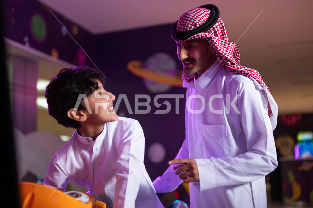 مفهوم الأبوة والبنوة، المشاركة في اللعب مع الأبناء، قضاء وقت ممتع مع العائلة، المراكز الترفيهيه المسلية في السعودية، صورة مقربة لولد عربي خليجي سعودي مع والده يقوم بركوب الدباب الكهربائي المفضل، الاستمتاع بألعاب الاركيد الالكترونية.