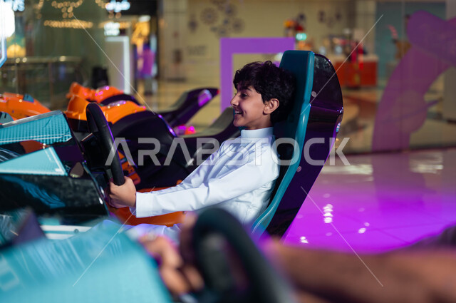 المتعة والتسلية في المراكز الترفيهية في السعودية، الشعور بالحماس والفرح، صورة من الجانب لولد عربي سعودي خليجي مبتسم يمسك المقود المتحرك يلعب لعبة محاكاة قيادة السيارات، ألعاب الأطفال الكهربائية، التركيز والقدرة على التحكم .