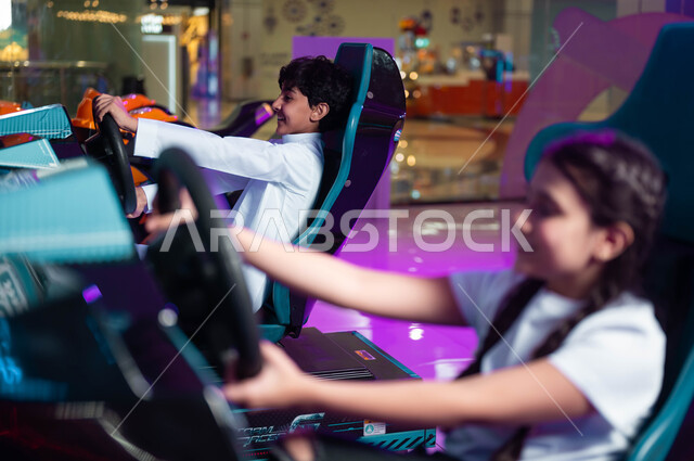 التركيز والقدرة على التحكم، التسلية والمتعة في المراكز الترفيهية في السعودية، الشعور بالحماس والفرح، صورة مقربة من الجانب لفتاة وولد عربيان سعوديان خليجيان مبتسمان يلعبان لعبة محاكاة قيادة السيارات، ألعاب الأطفال الكهربائية .