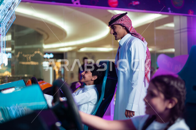 Participating in playing with children, focus and ability to control, the concept of fatherhood and sonship, a smiling Saudi Arabian Gulf father wearing a shemagh and traditional dress standing with his children in a car driving simulation game, children’s electric toys.