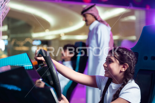 ألعاب الأطفال الكهربائية، المشاركة في اللعب مع الأبناء، التركيز والقدرة على التحكم، مفهوم الأبوة والبنوة، أب عربي خليجي سعودي مبتسم يرتدي الشماغ والثوب التقليدي يقف مع اولاده في لعبة محاكاة قيادة السيارات .