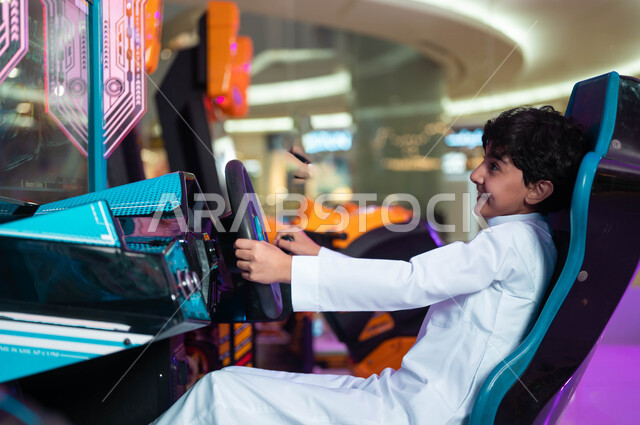 إيماءات الحماس والفرح، التركيز والقدرة على التحكم، ألعاب الأطفال الكهربائية، صورة من الجانب لولد عربي سعودي خليجي مبتسم يمسك المقود المتحرك يلعب في لعبة محاكاة قيادة السيارات، المراكز الترفيهية في المملكة العربية السعودية .