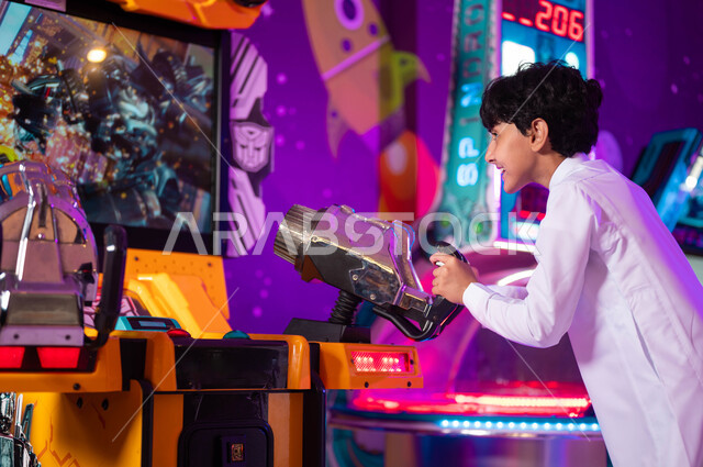 الاستمتاع بألعاب الأركيد المتطورة، ايماءات الحماس والفرح، قسم المنافسة والتحدي، الأماكن الترفيهية في السعودية، صورة من الجانب لولد عربي سعودي خليجي مبتسم يرتدي الثوب التقليدي يلعب لعبة إطلاق النار الالكترونية، التركيز والقدرة على التحكم  .