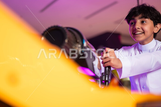 التركيز لإصابة الهدف، الاستمتاع بألعاب الأركيد الحديثة، ايماءات الحماس والفرح، قسم المنافسة والتحدي، الأماكن الترفيهية في السعودية، صورة مقربة لولد عربي سعودي خليجي مبتسم يرتدي الثوب التقليدي يلعب لعبة إطلاق النار الالكترونية .