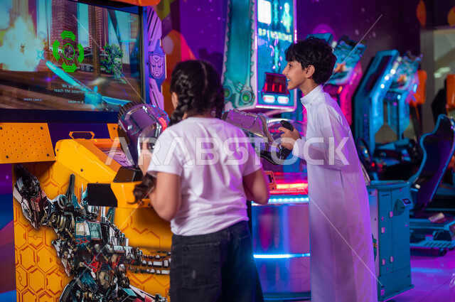 التنافس في اللعب بين الأخوة، التركيز لإصابة الهدف، الاستمتاع بألعاب الأركيد الحديثة و المتنوعة، قسم المنافسة والتحدي، صورة من الخلف لولد وفتاة عربيان سعوديان خليجيان يلعبان لعبة إطلاق النار الالكترونية، الأماكن الترفيهية في السعودية .