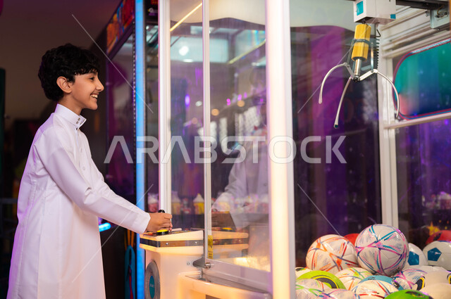 الشعور بالمتعة والفرح، المراكز الترفيهية في المملكة العربية السعودية، التركيز والقدرة على التحكم، صالات ألعاب الأركيد المتطورة، صورة من الجانب لولد عربي سعودي خليجي يرتدي الثوب التقليدي يلعب آلة مخلب الالتقاط الالكترونية، قسم التحديات والمغامرات.