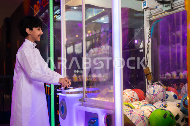 قسم التحديات والمغامرات، صالات ألعاب الأركيد الالكترونية، الاستمتاع بالأنشطة الترفيهية المتميزة، التركيز والقدرة على التحكم، صورة من الجانب لولد عربي سعودي خليجي يرتدي الثوب التقليدي يلعب آلة مخلب الالتقاط، قضاء إجازة صيفية ممتعة .