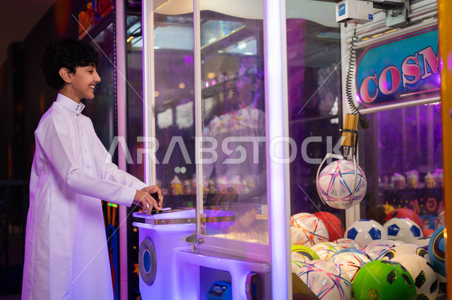 صالات ألعاب الأركيد المتطورة، الاستمتاع بالأنشطة الترفيهية المتميزة، التركيز والقدرة على التحكم، قسم التحديات والمغامرات، صورة من الجانب لولد عربي سعودي خليجي يرتدي الثوب التقليدي يلعب آلة مخلب الالتقاط الالكترونية، قضاء إجازة صيفية ممتعة .