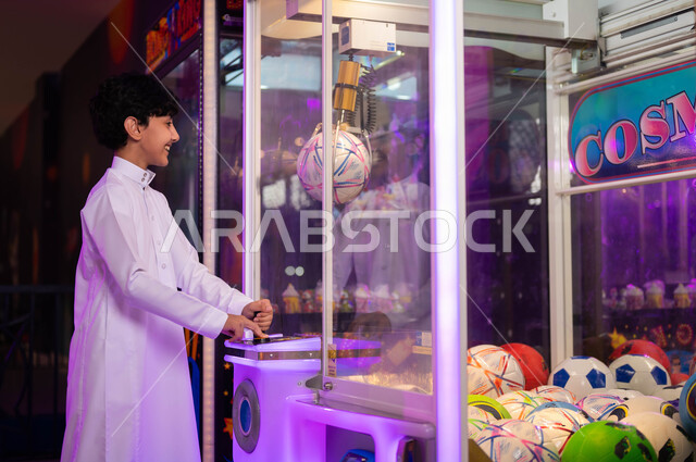 Concentration and ability to control, entertainment centers in the Kingdom of Saudi Arabia, electronic arcade games halls, challenges and adventures section, a picture from the side of a Saudi Arabian Gulf boy wearing a traditional dress playing an electronic pick-up claw machine, spending a fun summer vacation
