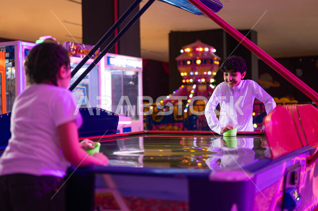 Enjoying sliding disc competitions and matches between brothers, distinct entertainment activities, electronic arcade games halls, challenges and adventures section, two Saudi Gulf Arab boys and girls competing in air hockey games on the electronic table