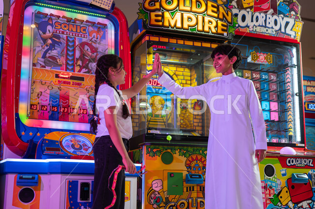 مشاركة المغامرات والتحديات بين الأخوة، الشعور بالحماس والفرح، المراكز الترفيهية في المملكة العربية السعودية، ألعاب الأركيد الالكترونية، فتاة عربية سعودية خليجية تلعب مع أخيها يرفعان يديهما بإيماءات الفوز والربح.