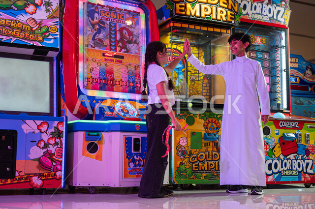 الاستمتاع في اللعب والتحدي بين الأخوة، الشعور بالحماس والفرح، المراكز الترفيهية في المملكة العربية السعودية، ألعاب الأركيد الالكترونية، فتاة عربية سعودية خليجية تلعب مع أخاها يرفعان يديهما بإيماءات الفوز والربح، صورة الجسم بالطول الكامل.