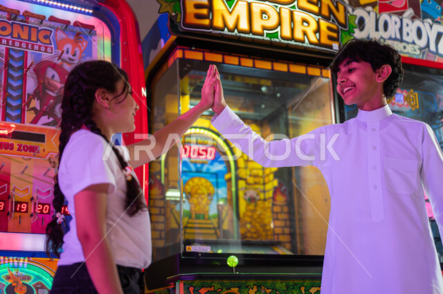 الاستمتاع في اللعب والمشاركة مع الأخوة، الشعور بالحماس والفرح، المراكز الترفيهية في المملكة العربية السعودية، ألعاب الأركيد الالكترونية، صورة مقربة لفتاة عربية سعودية خليجية تلعب مع أخاها يرفعان يديهما بإيماءات الفوز والربح .