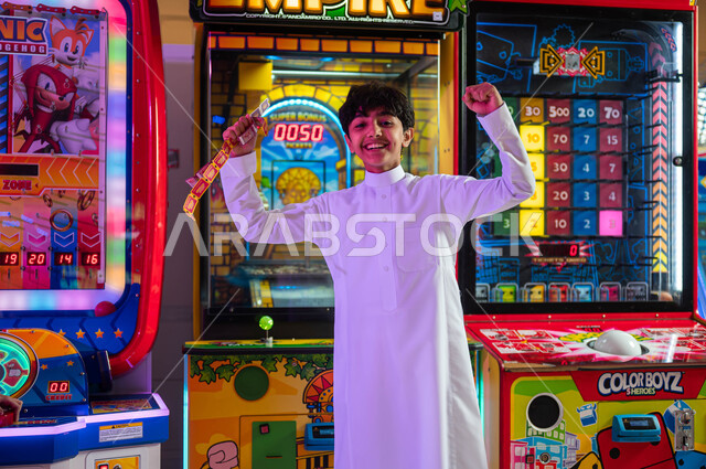 Feeling joy and happiness, entertainment centers in the Kingdom of Saudi Arabia, spending fun and entertaining times, a Saudi Gulf Arab boy wearing a traditional dress, looking at the camera, raising his hands with win-win gestures, enjoying the summer vacation.
